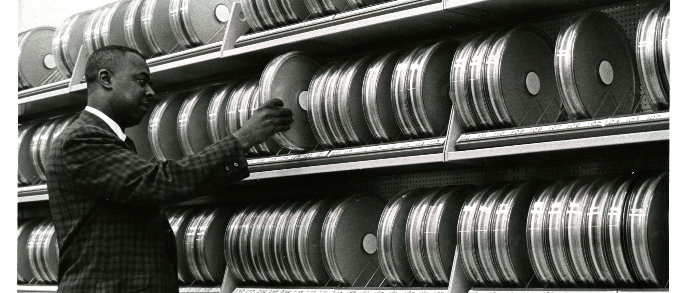 An archivist drawing one numbered canister from a wall of indexed shelving.